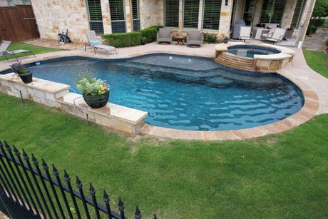 Backyard pool with curved stone deck, hot tub, lawn, and patio near a house