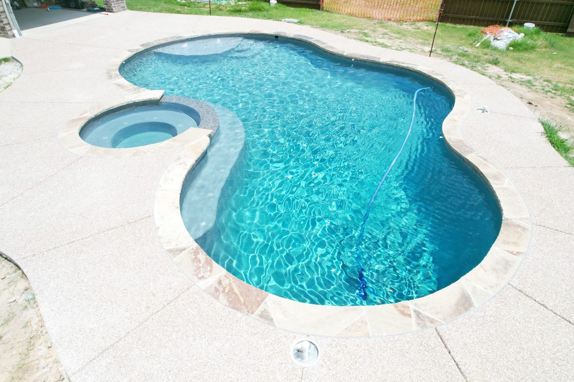Kidney-shaped backyard pool with turquoise water and an attached spa, surrounded by light stone decking.