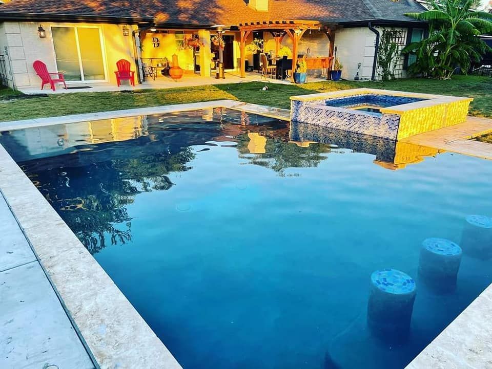 Backyard pool beside a house, with warm patio lights and a stone water feature at dusk