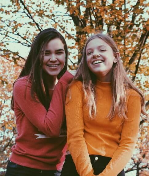Twee lachende vrienden buiten in de herfst, gekleed in rode en oranje truien onder een boom.