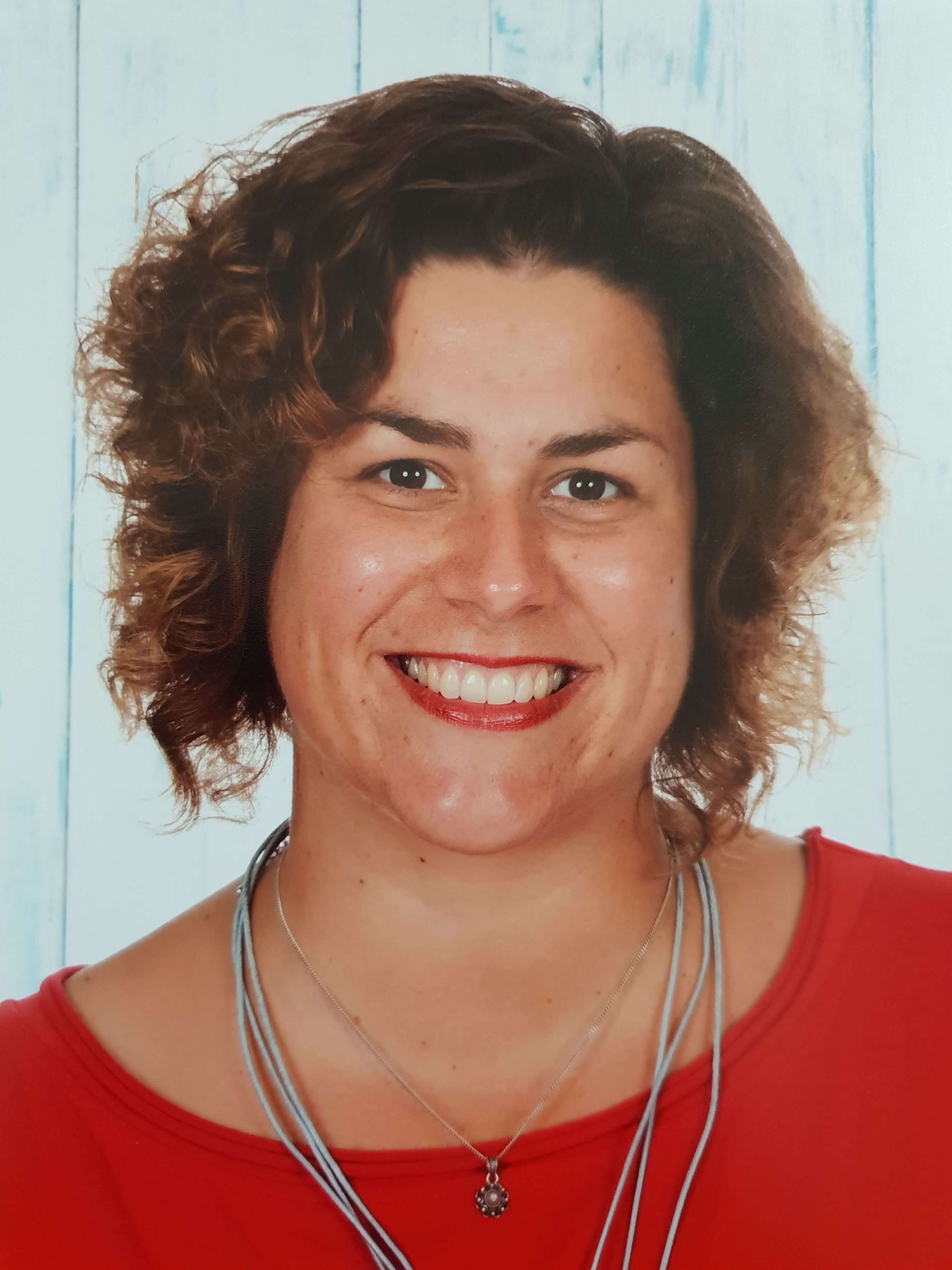 Lachende vrouw met krullend bruin haar, gekleed in een rode top en meerdere kettingen, tegen een lichte achtergrond.