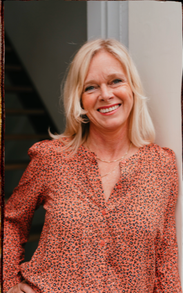 Lachende blonde vrouw in een rode blouse met patroon staat binnen bij een trap.
