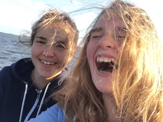 Twee lachende mensen buiten bij het water, met wapperend haar in de wind, en één van hen lacht.