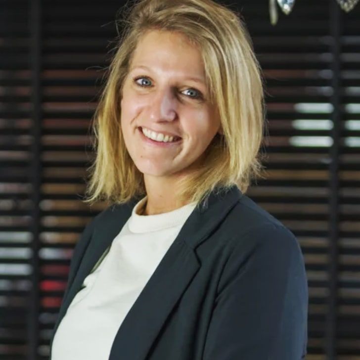 Een lachend persoon in een donkere blazer en witte top voor een donker raam met jaloezieën.