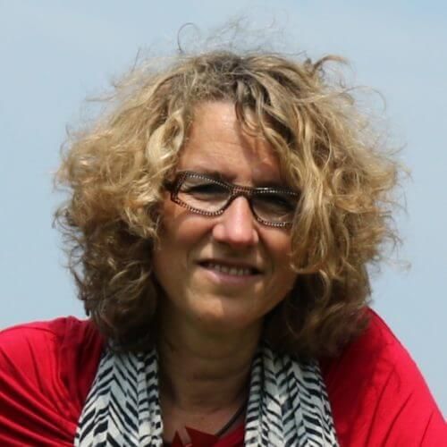 Vrouw met krullend blond haar en een bril, gekleed in een rode top en een gestreepte sjaal, lacht in de buitenlucht.