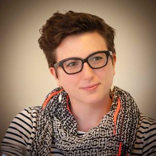 Portret van een persoon met een bril en een sjaal met patroon, die zachtjes glimlacht tegen een neutrale achtergrond.
