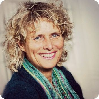 Een lachend persoon met krullend blond haar en een turquoise sjaal, tegen een neutrale achtergrond.