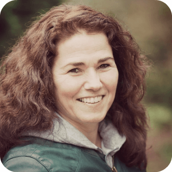Lachende vrouw in een groene jas met krullend bruin haar, staand buiten tegen een wazige achtergrond.