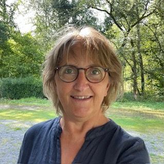 Een lachend persoon met een bril en een donkerblauw shirt bevindt zich buiten tussen groene bomen.