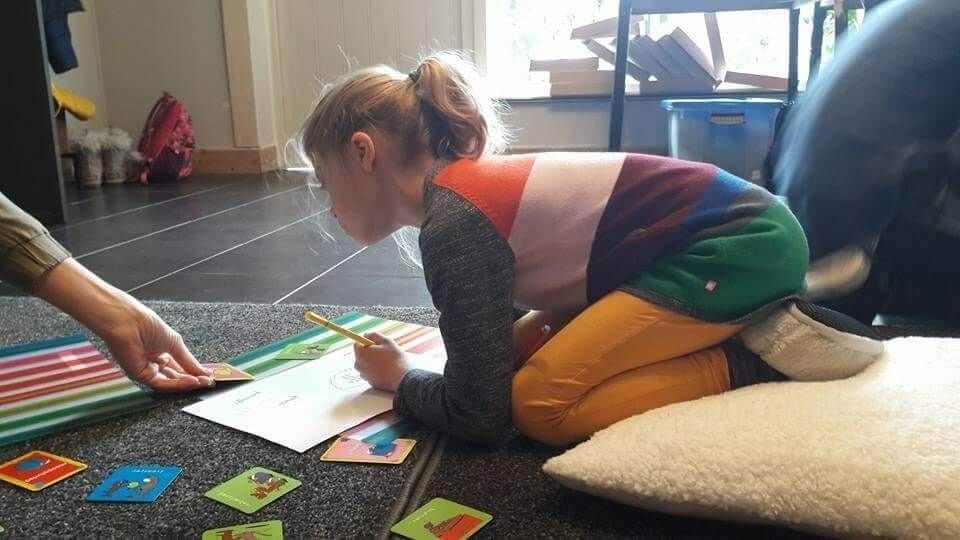 Een kind knielt op een mat en reikt naar kleurrijke kaarten, terwijl een volwassene in de buurt in een speelkamer wijst.