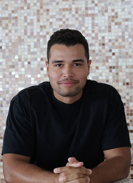 Man with dark hair and a black shirt, smiling, arms crossed. Mosaic tile background.