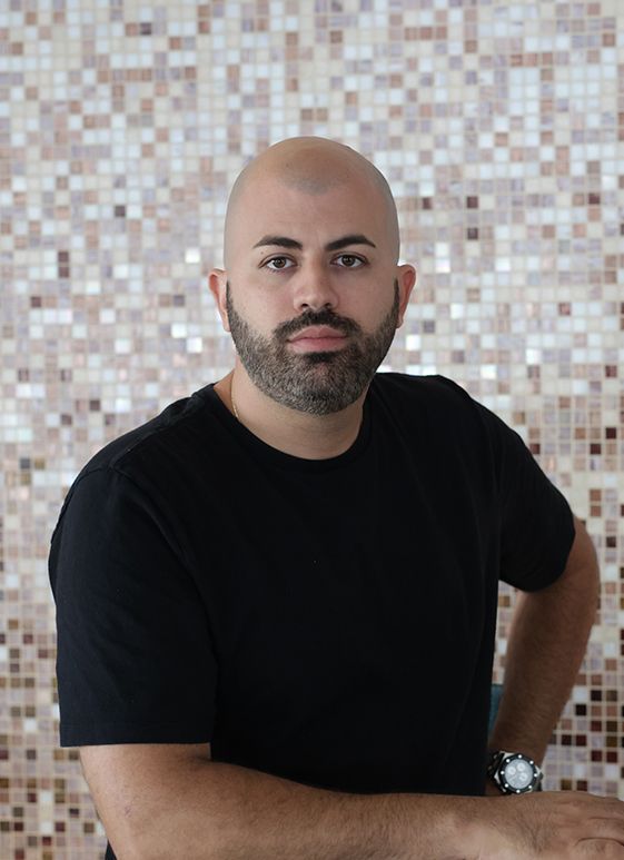 Bald man with a beard in a black shirt, leaning against a mosaic-tiled wall.