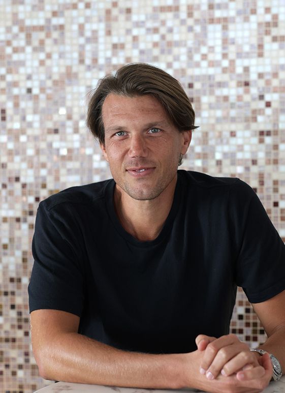 Man in black t-shirt smiles, arms crossed, in front of a mosaic tile wall.