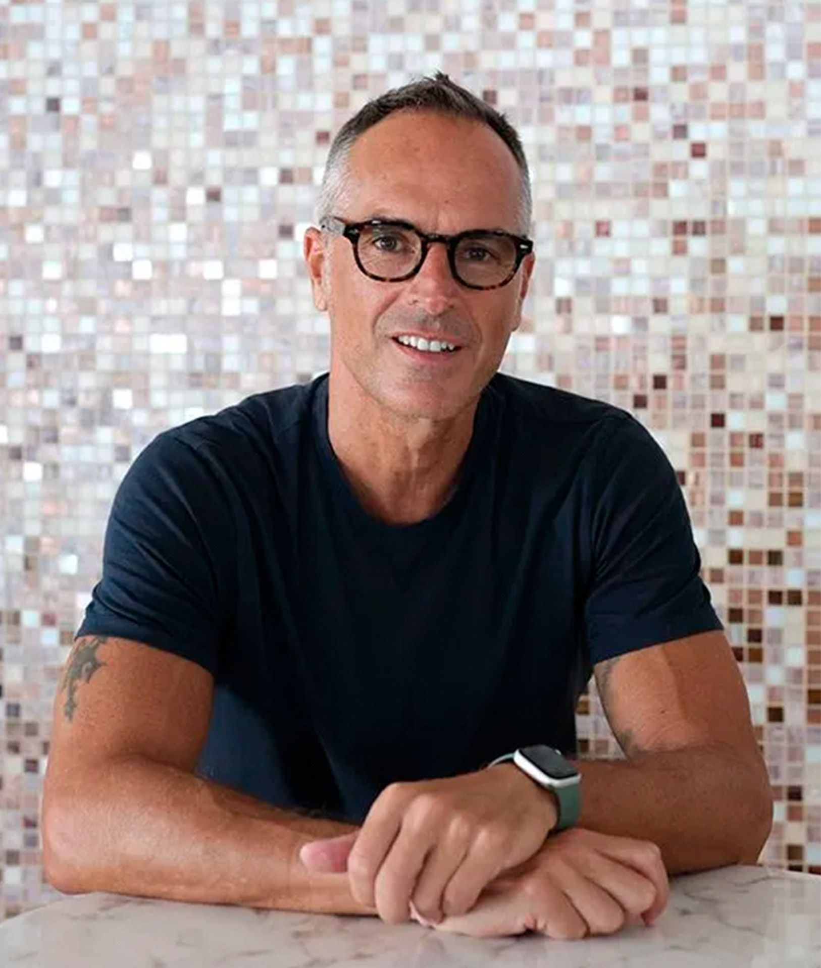 Man with glasses, arms crossed, smiling, in front of mosaic tile wall. Wearing a dark blue shirt and watch.