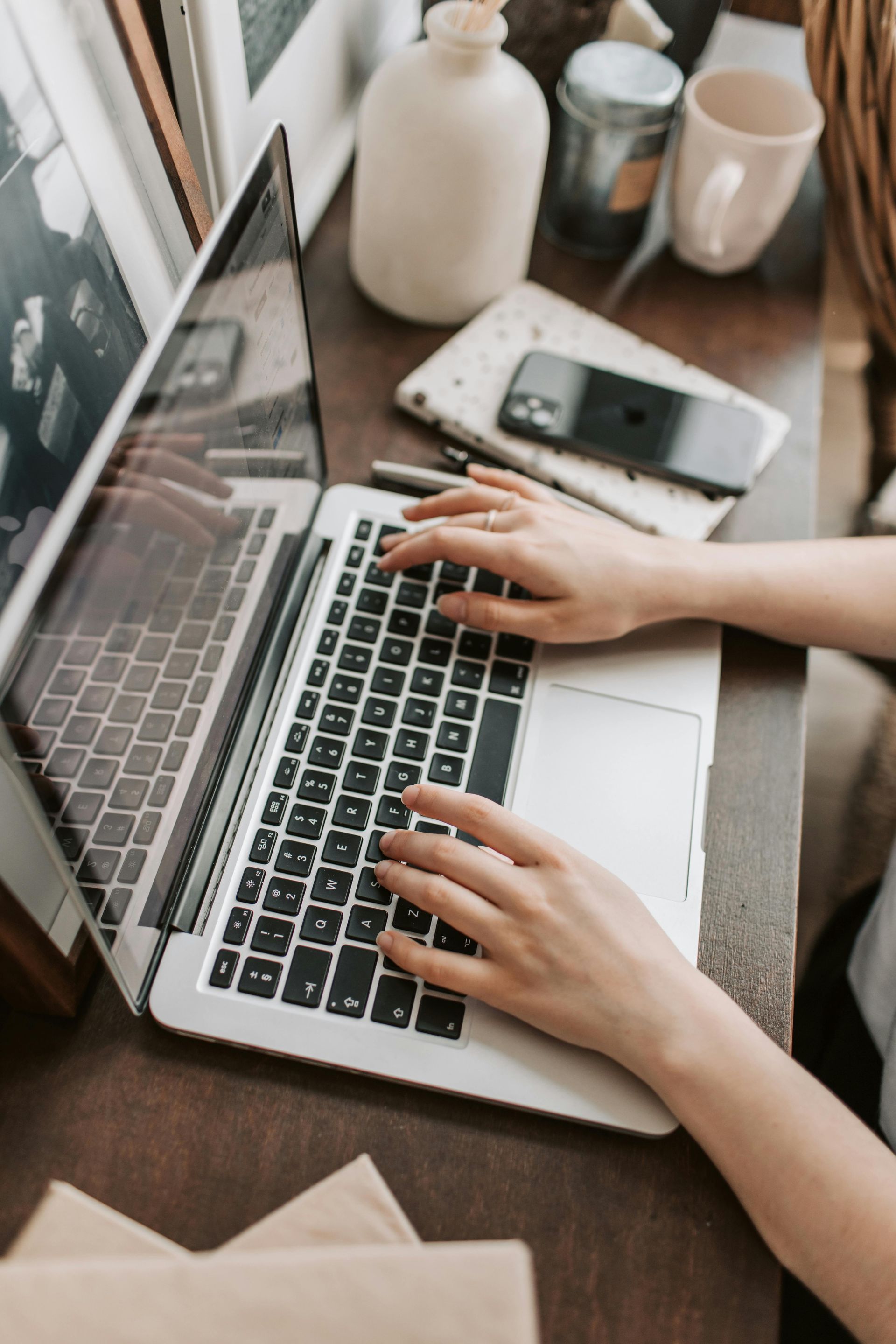 Hands typing on a laptop keyboard at a table with a coffee cup; two people in suits blurred in the background.