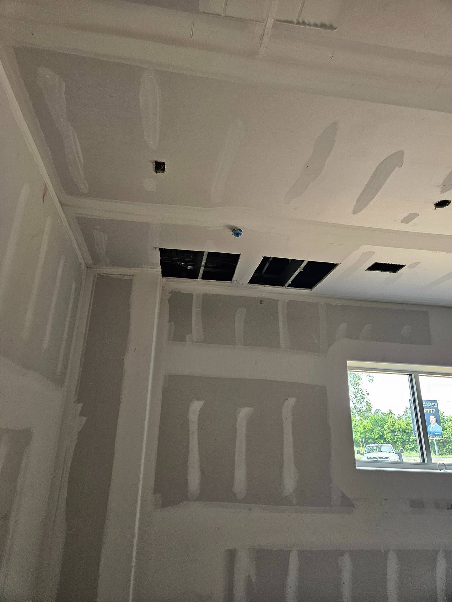 Unfinished drywall room with ceiling cutouts and a window showing greenery outside