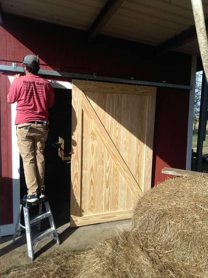 Person on a ladder installing a wooden barn door on a red shed, with hay piled nearby.