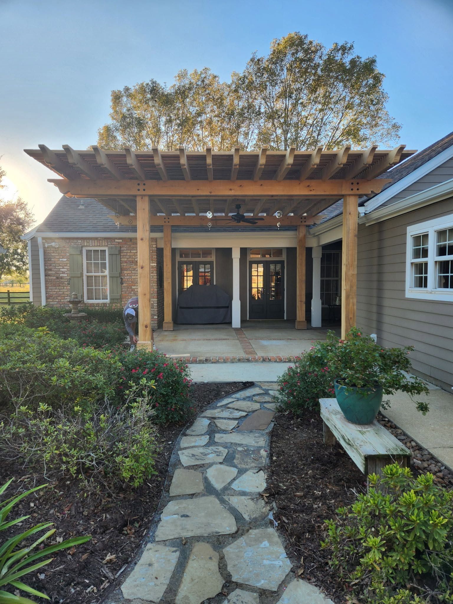 Stone path leading to a house entrance with wooden pergola, double doors, and landscaped shrubs.