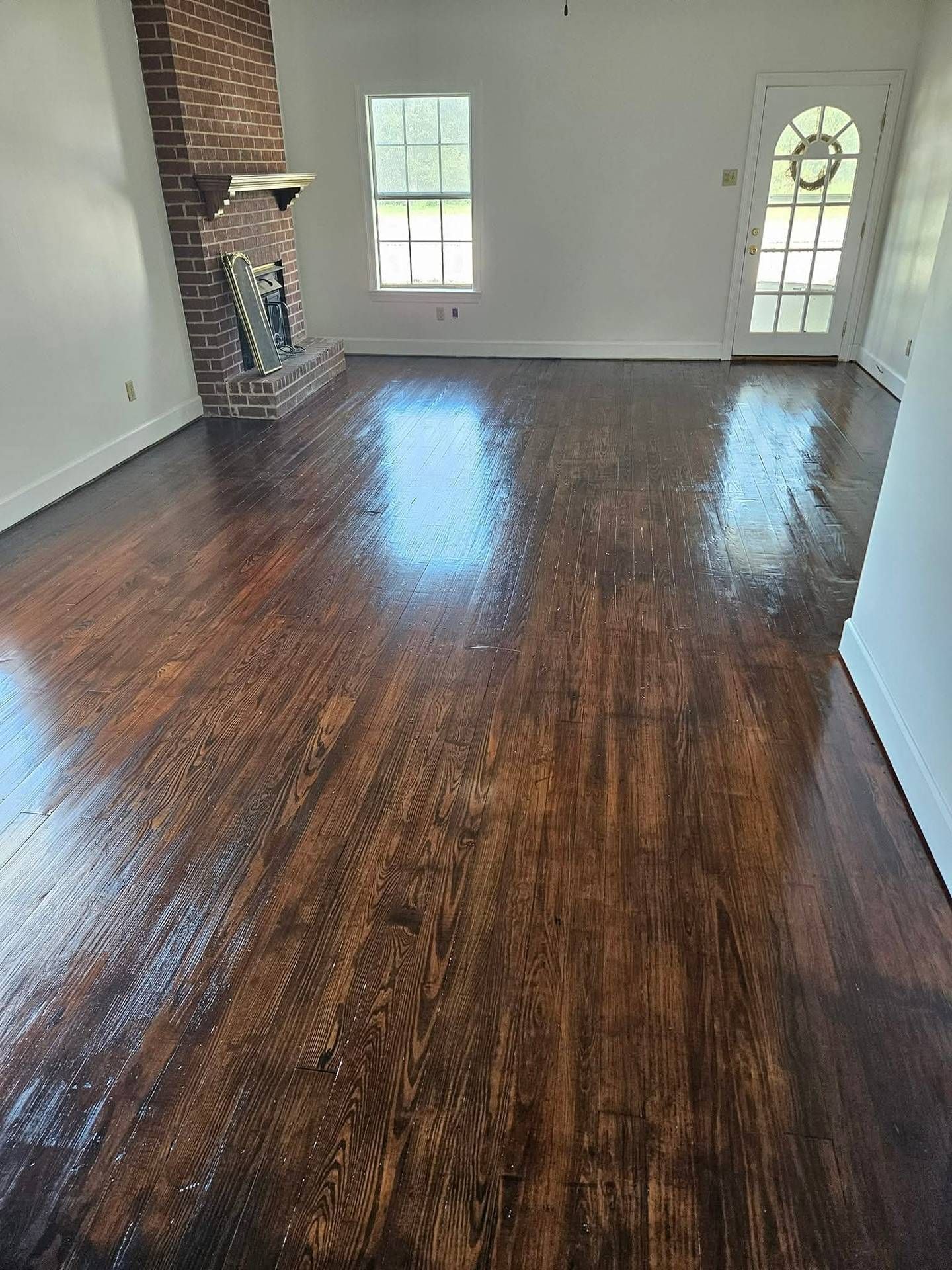 Empty living room with glossy dark hardwood floors, white walls, brick fireplace, and two windows.