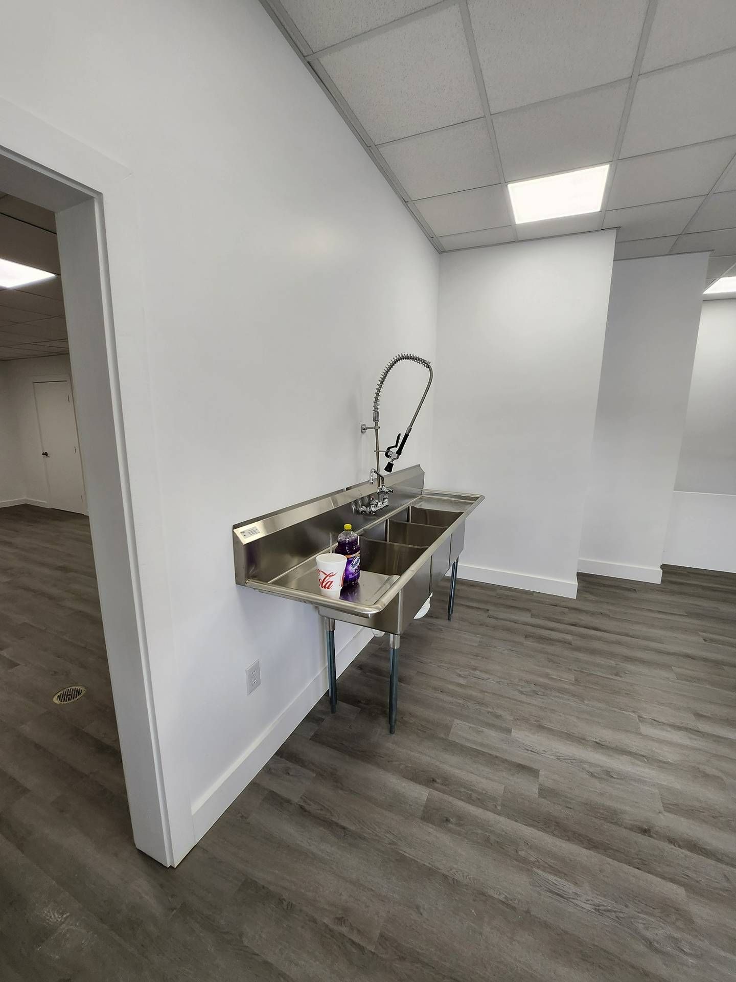Empty break room with stainless sink, white walls, gray wood floor, and a purple cleaning bottle.