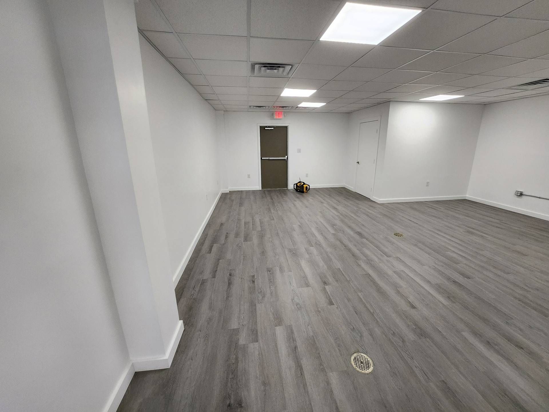Empty office room with gray carpet, white walls, ceiling lights, and a door at the back.