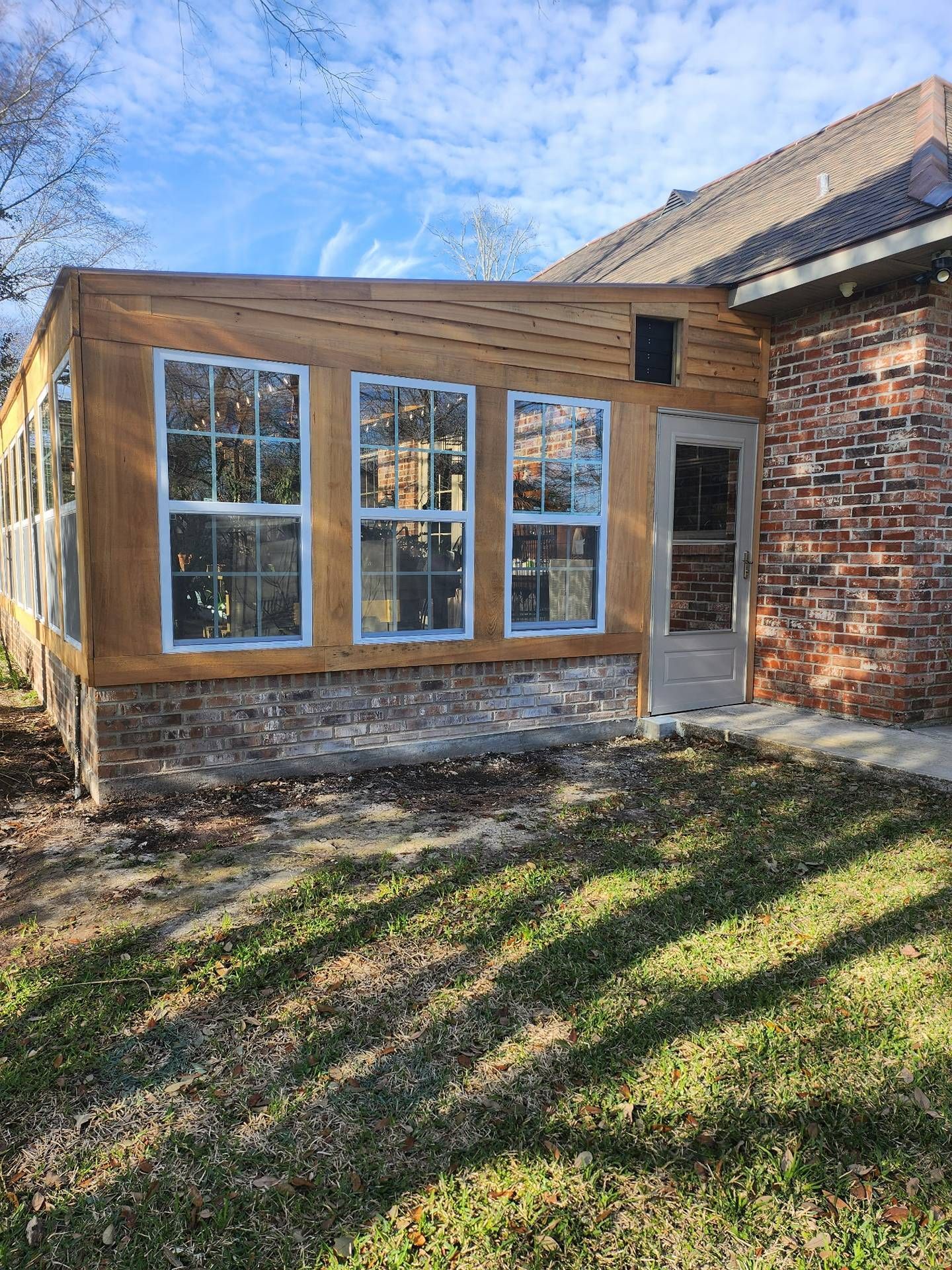 Small brick and wood house addition with large windows and a side door beside a grassy yard.