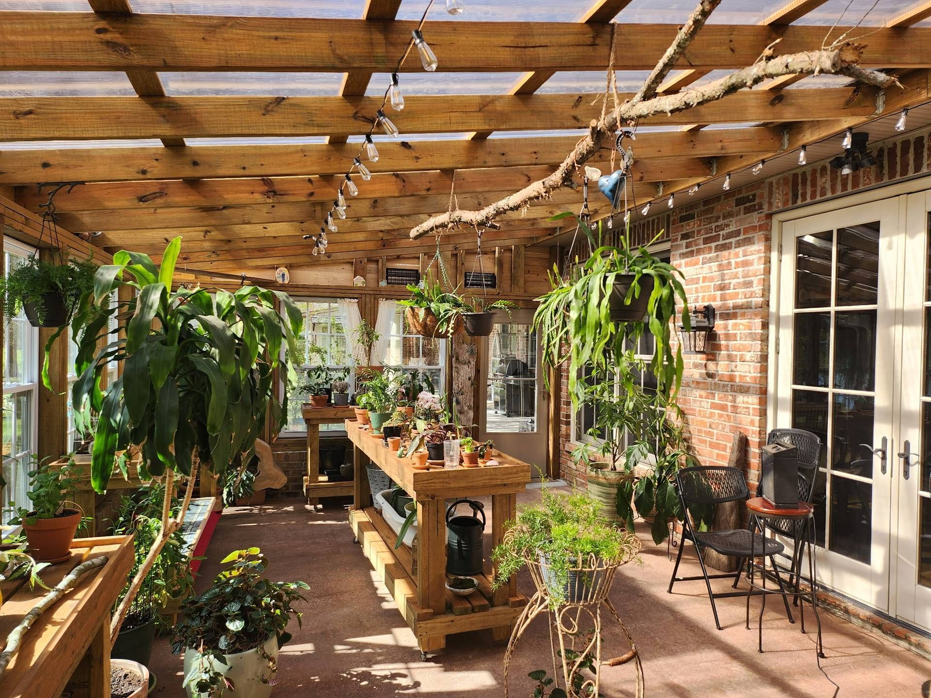 Sunlit greenhouse patio with hanging plants, wooden tables, and brick walls beside a glass door