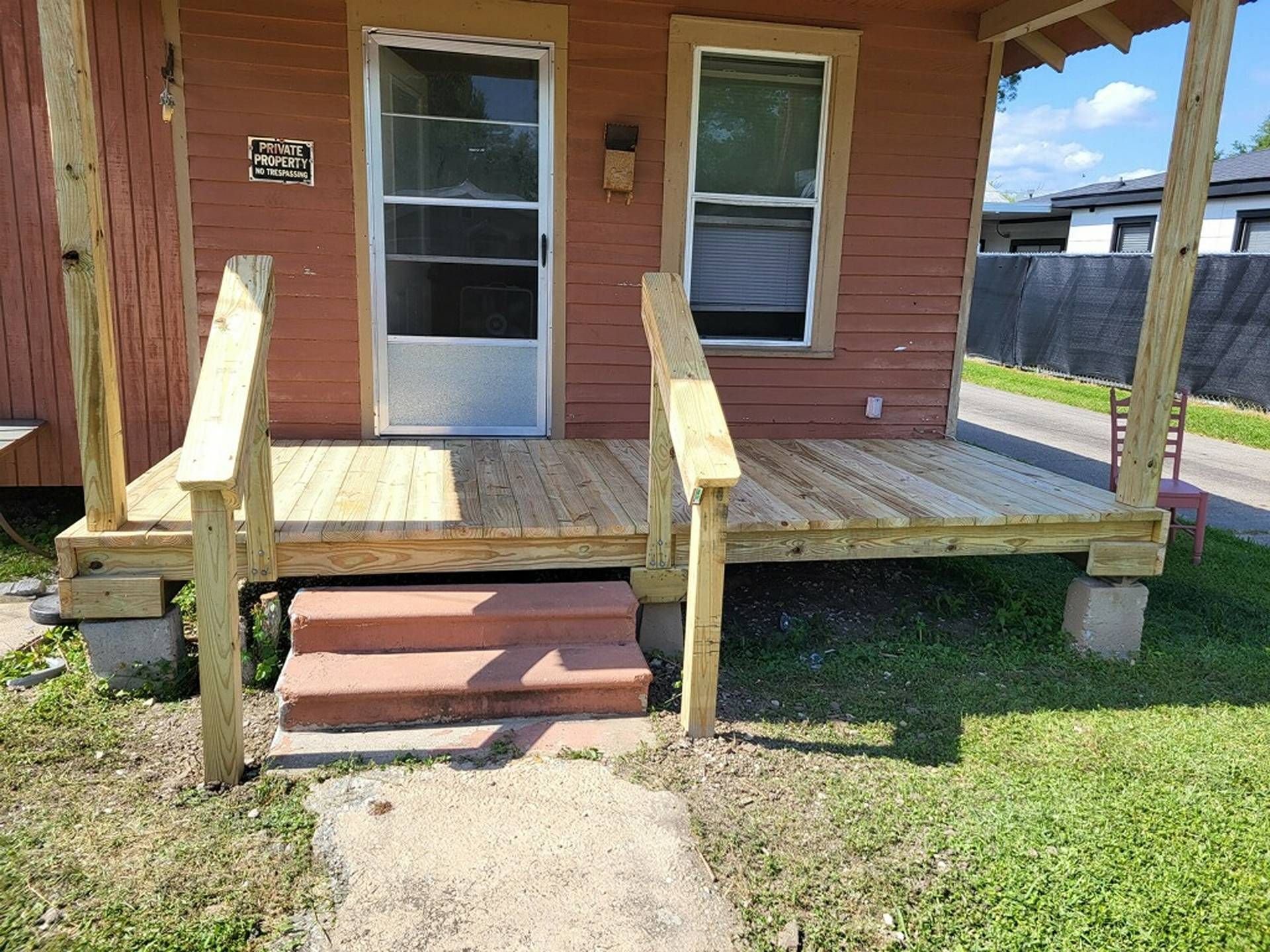 Small wooden front porch with steps and railings on a brick house, beside a grassy yard.