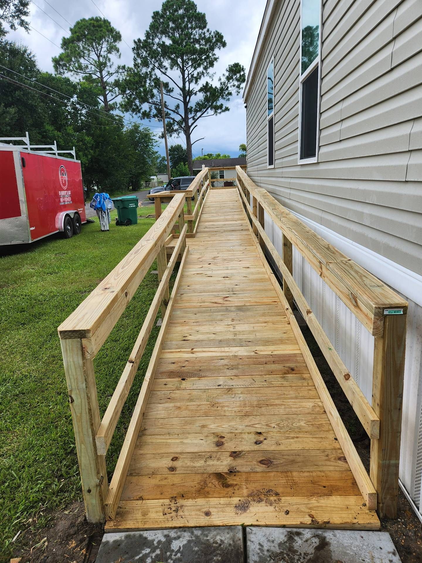 Wooden accessible ramp beside a house, with handrails and a red container in the yard.
