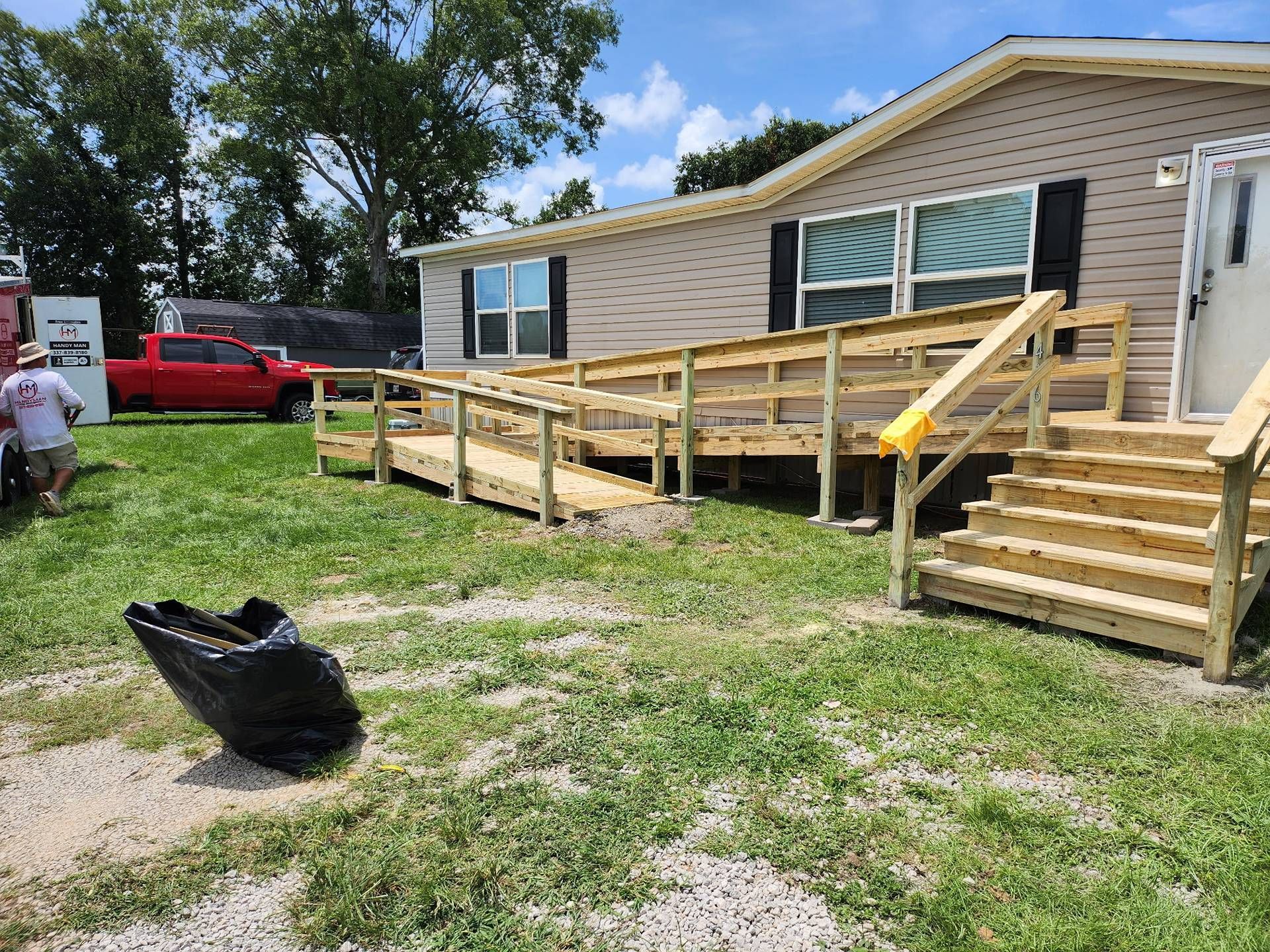 House with wooden ramp and stairs in a yard, with a black trash bag in the foreground and a red truck nearby