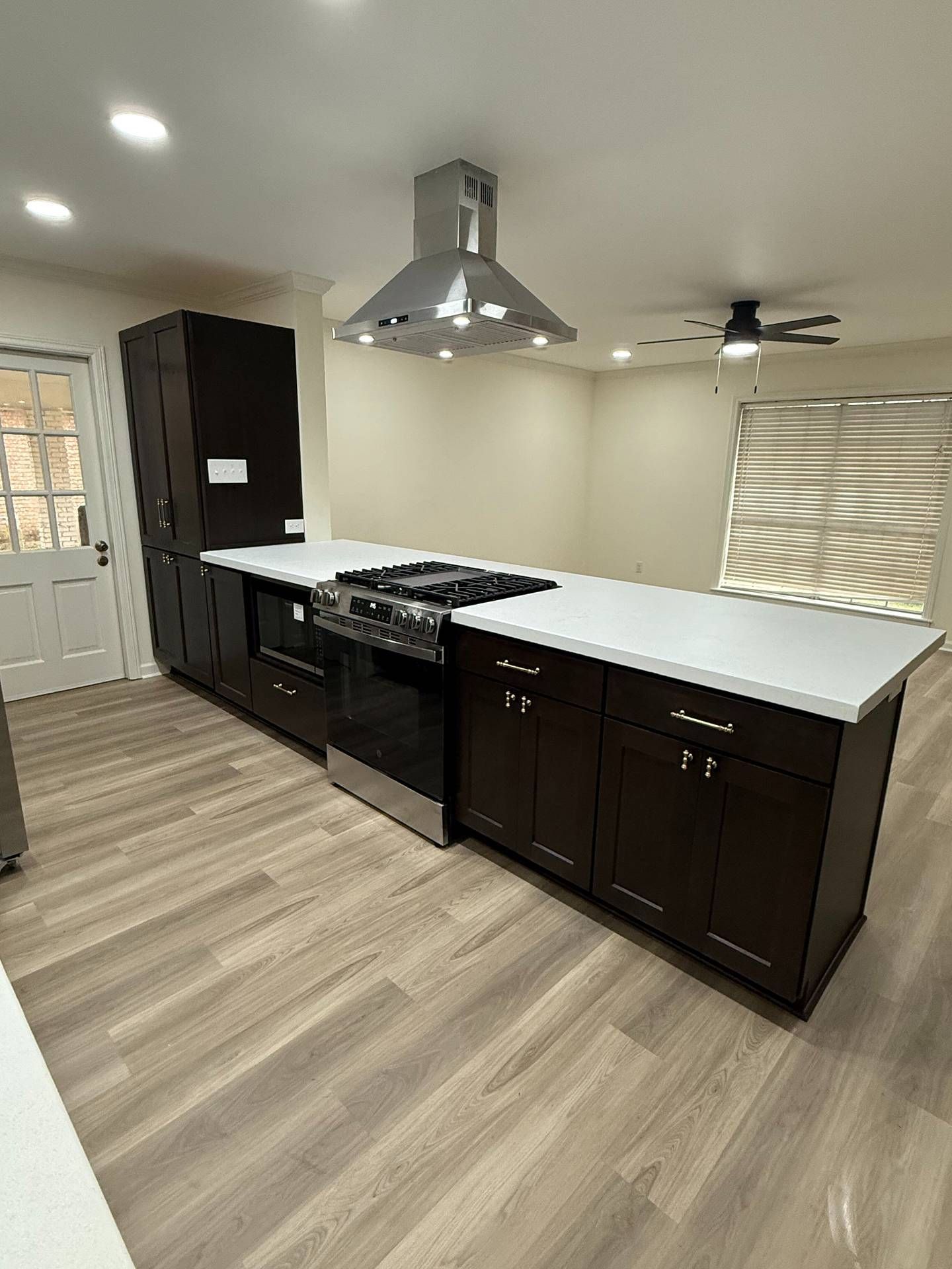 Modern kitchen with dark cabinets, white island counters, stainless range hood, and wood-look flooring