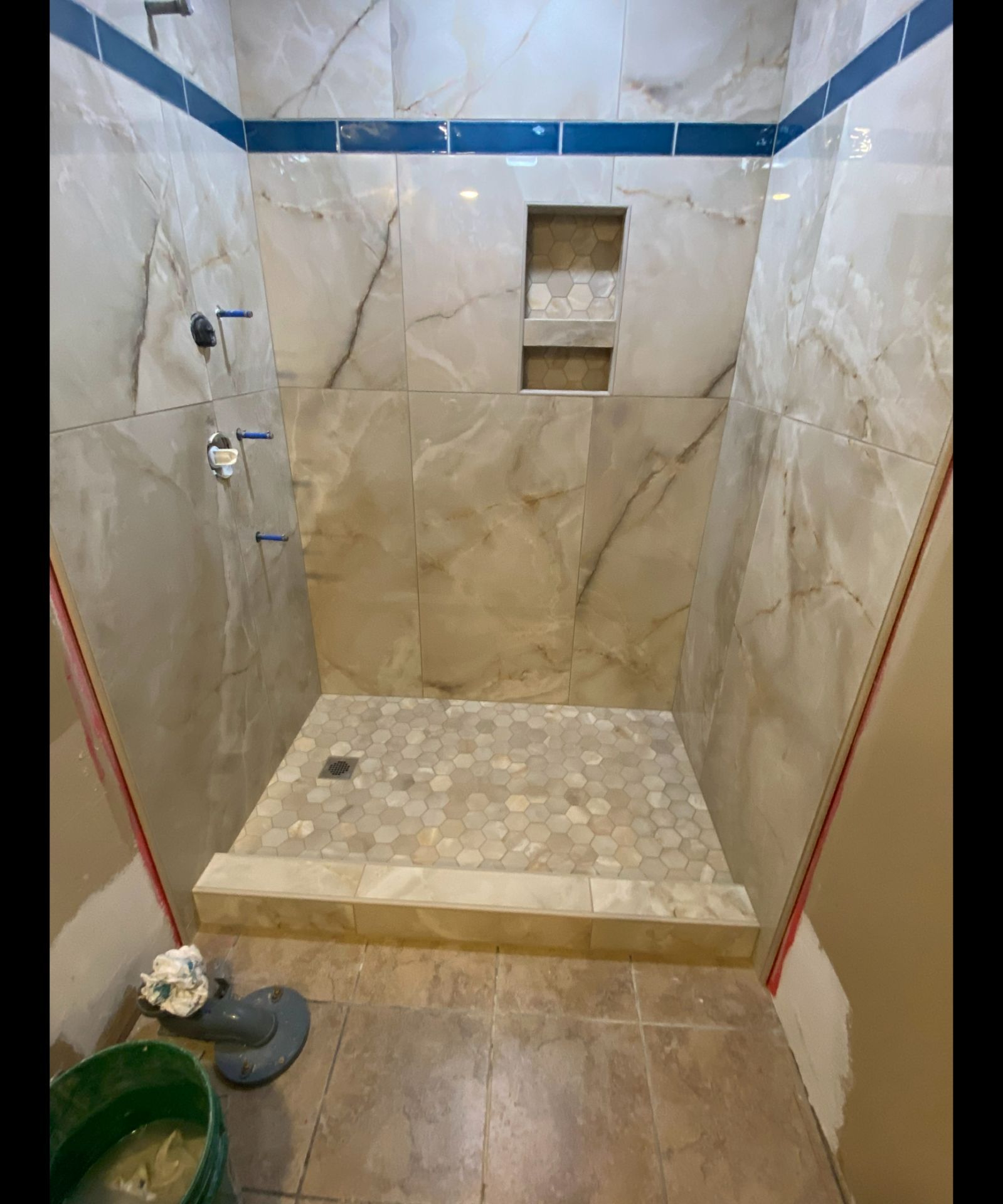 Empty tiled shower with beige marble walls, blue accent strip, and a small recessed shelf.