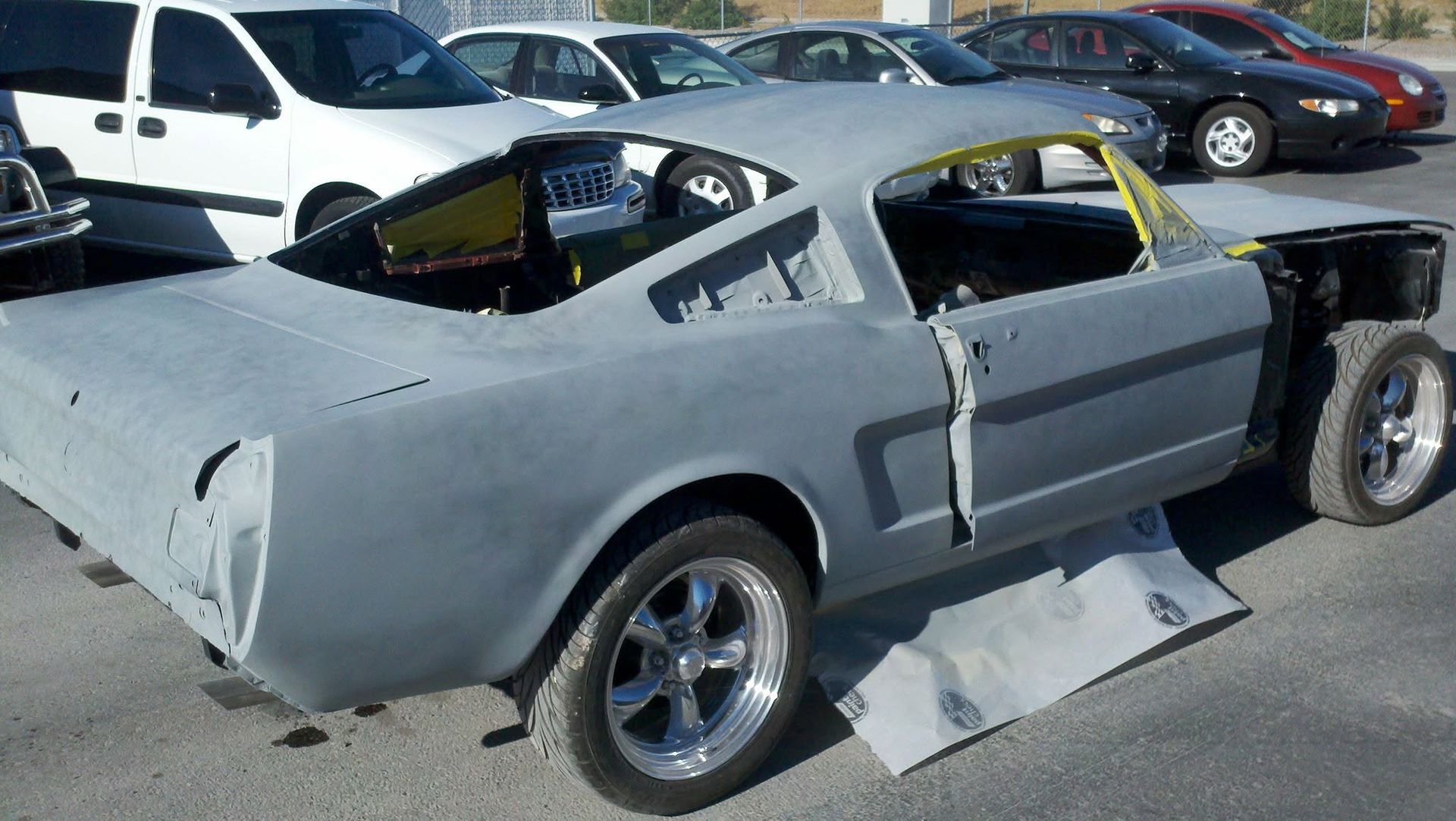 Gray classic car body shell parked in a lot, missing front end and windows, with detached parts on the ground