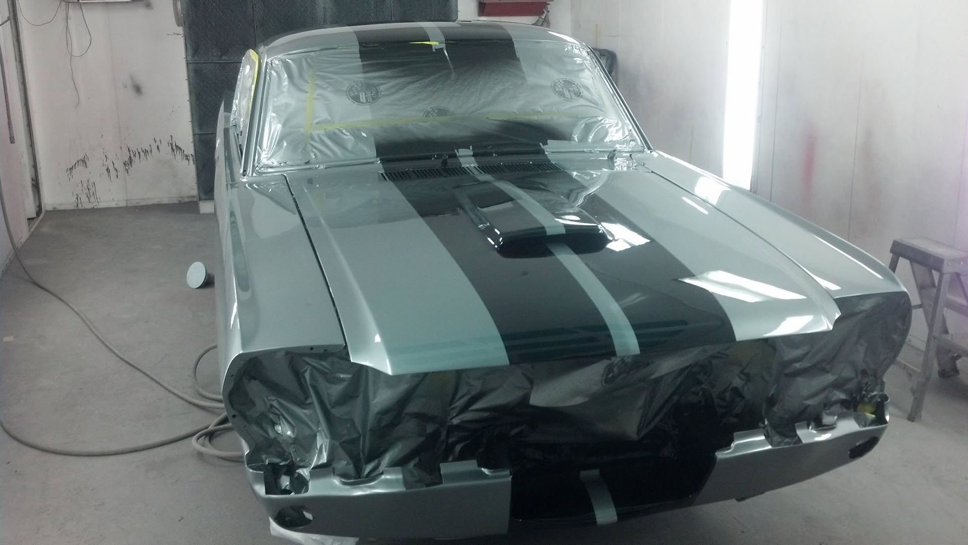 Classic car in a paint booth, mostly gray with black racing stripes and covered headlights.