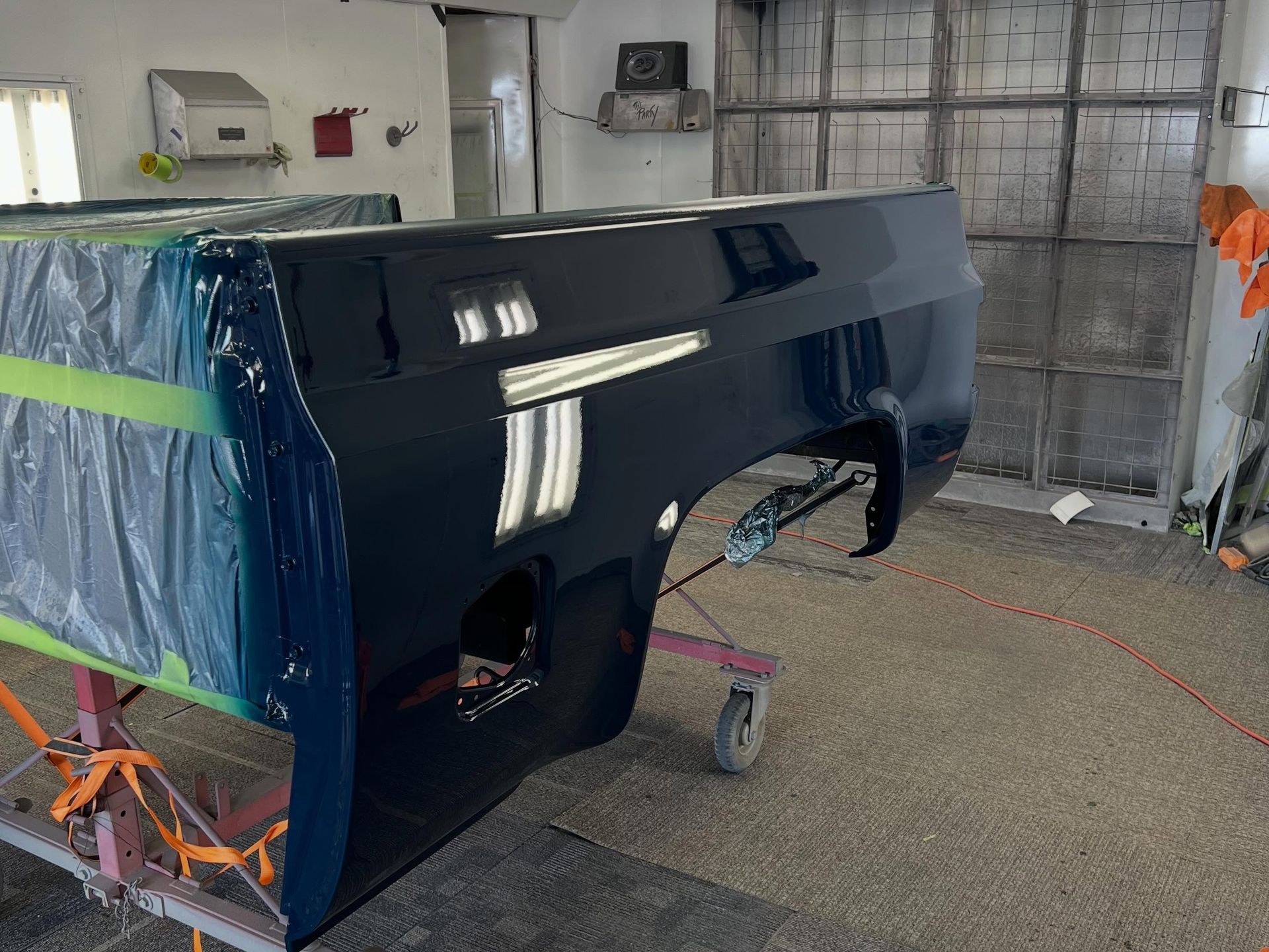 Glossy dark blue car body panel in a workshop, mounted on a stand and covered with masking tape and plastic.