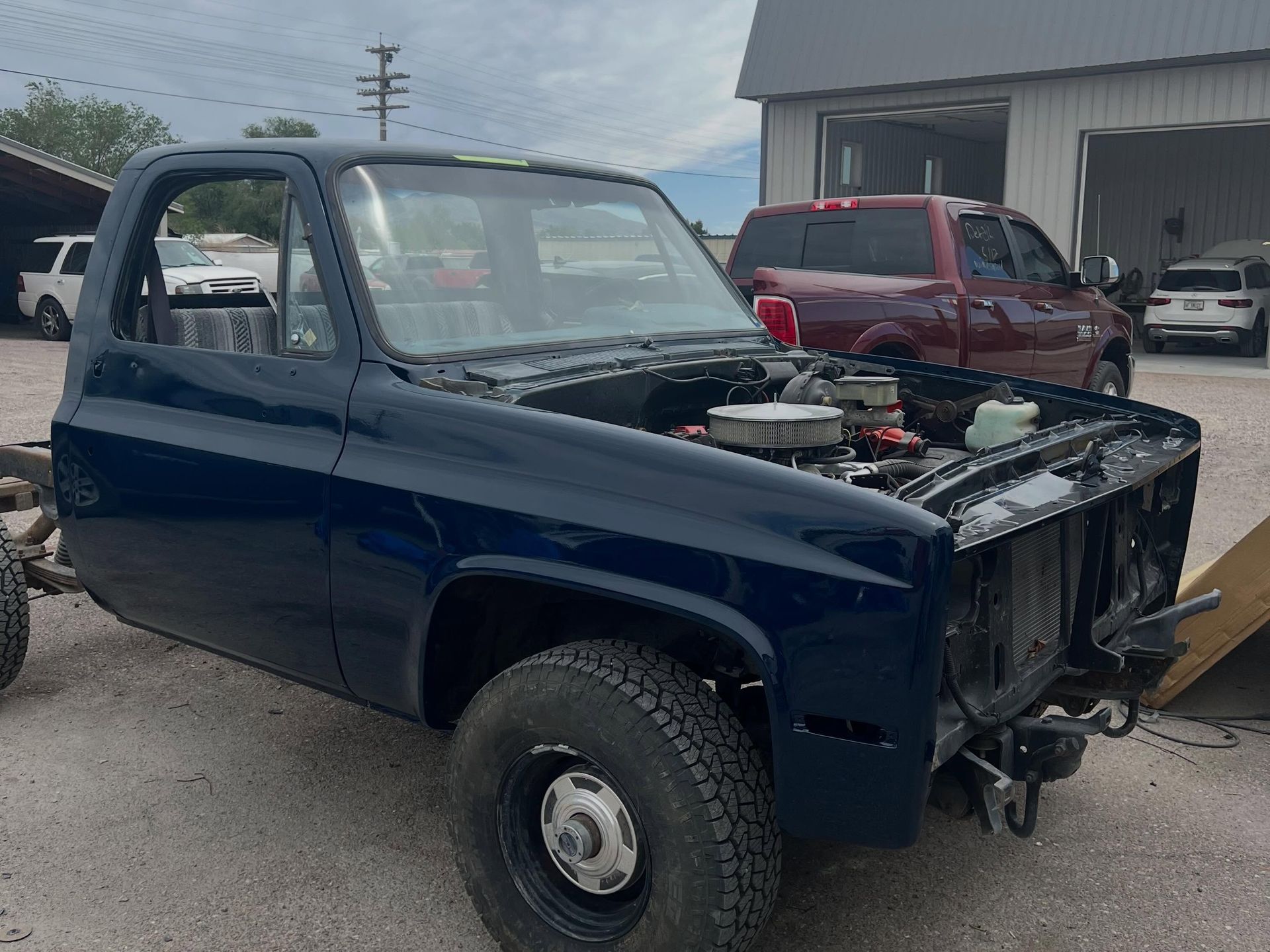 Blue pickup truck cab and front end stripped, parked in a lot with a red truck and warehouse in the background