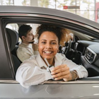 Smiling driver at a car dealership, holding keys with passenger seated inside a silver car