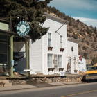 Small-town street with a white storefront and clock sign, parked yellow pickup, and hillside in the background.