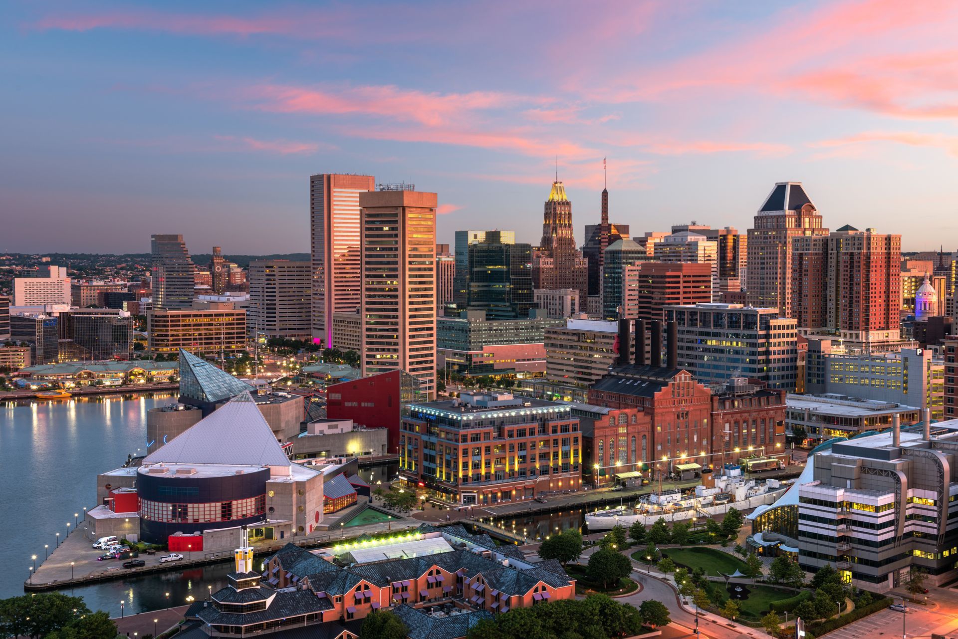 Baltimore skyline at dusk with colorful sky reflected in the water. Buildings lit up.