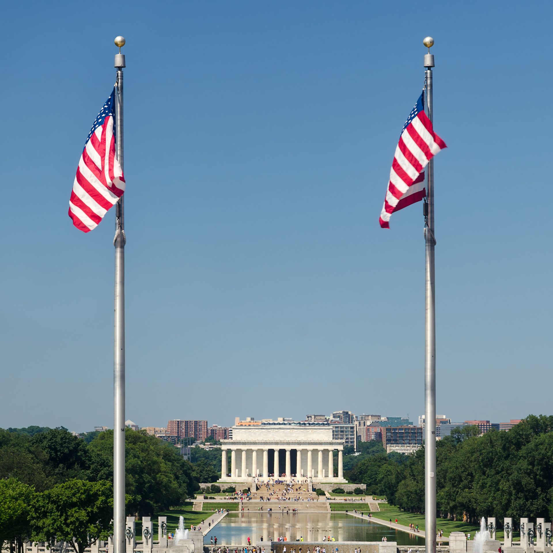 Two American flags flank the Lincoln Memorial in Washington, D.C., reflecting in a long pool, clear blue sky.
