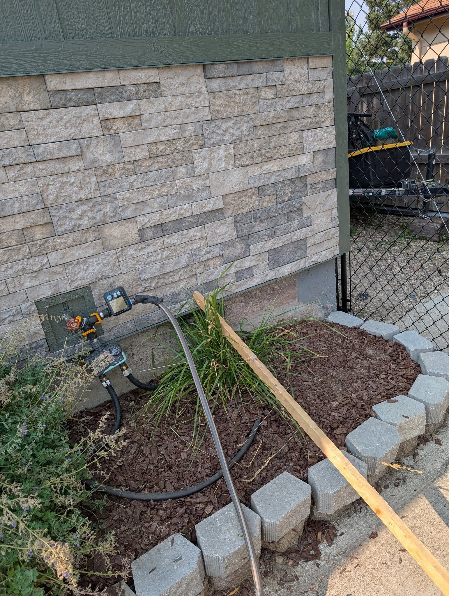 Garden hose spraying water onto a stucco wall beside a small landscaped yard and stone border.