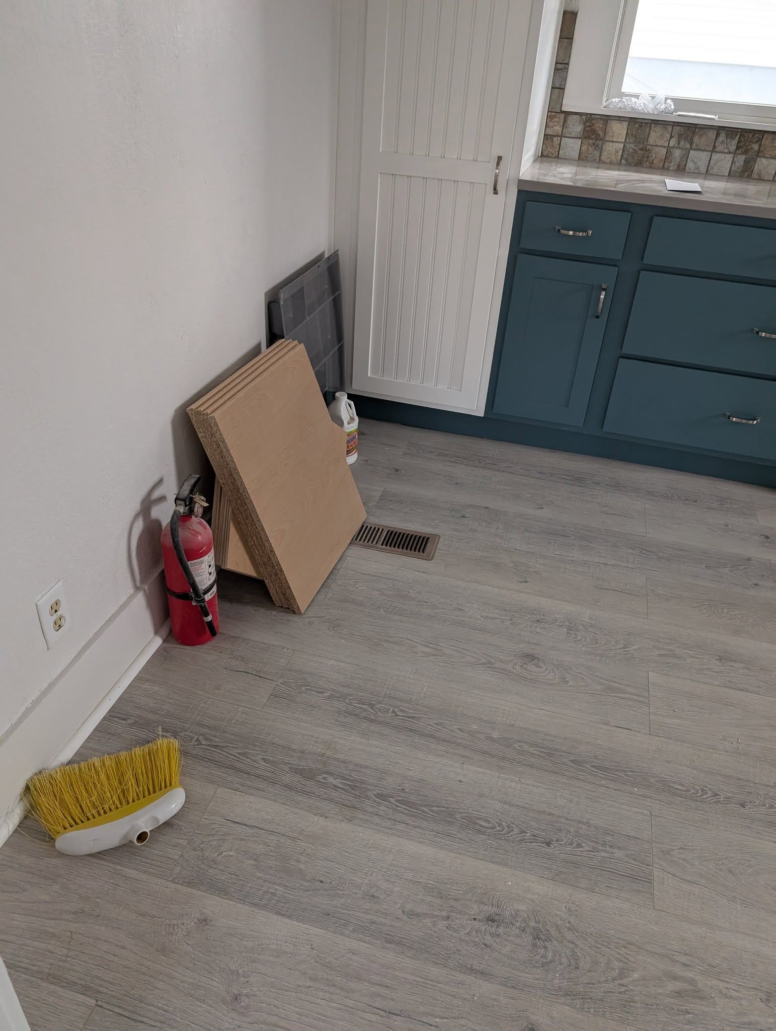 Kitchen corner with a fire extinguisher, leaning cardboard, and a small brush on the floor beside teal cabinets.