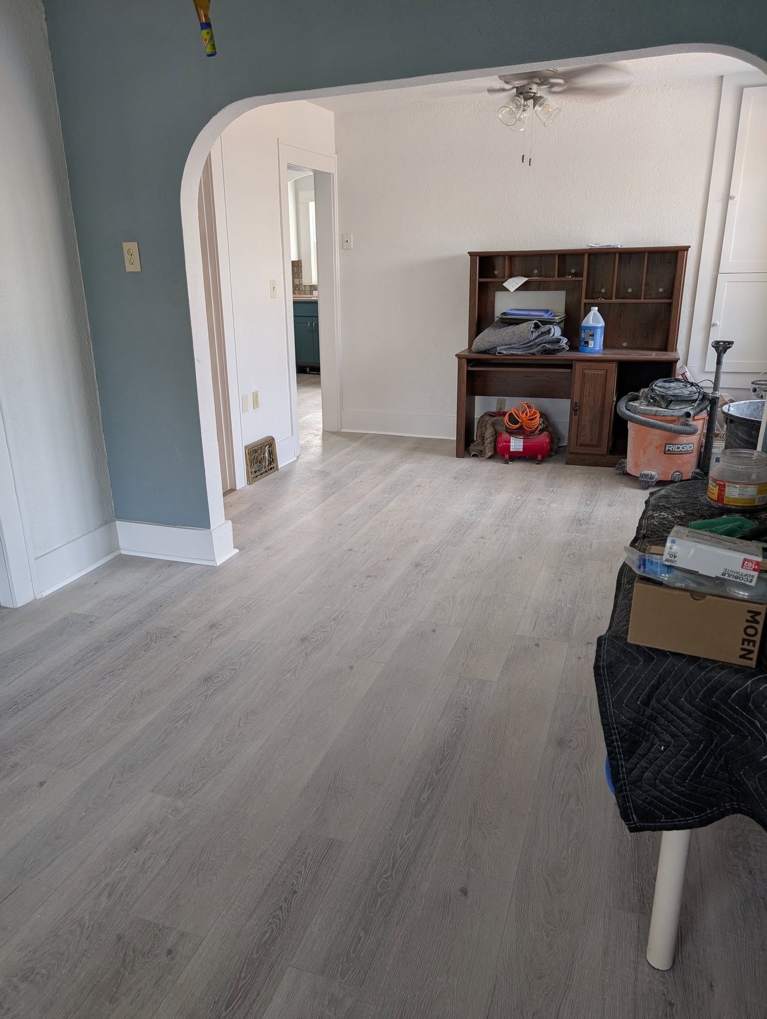 Empty room with light wood floor, white archway, and a shelf with assorted items on the right.