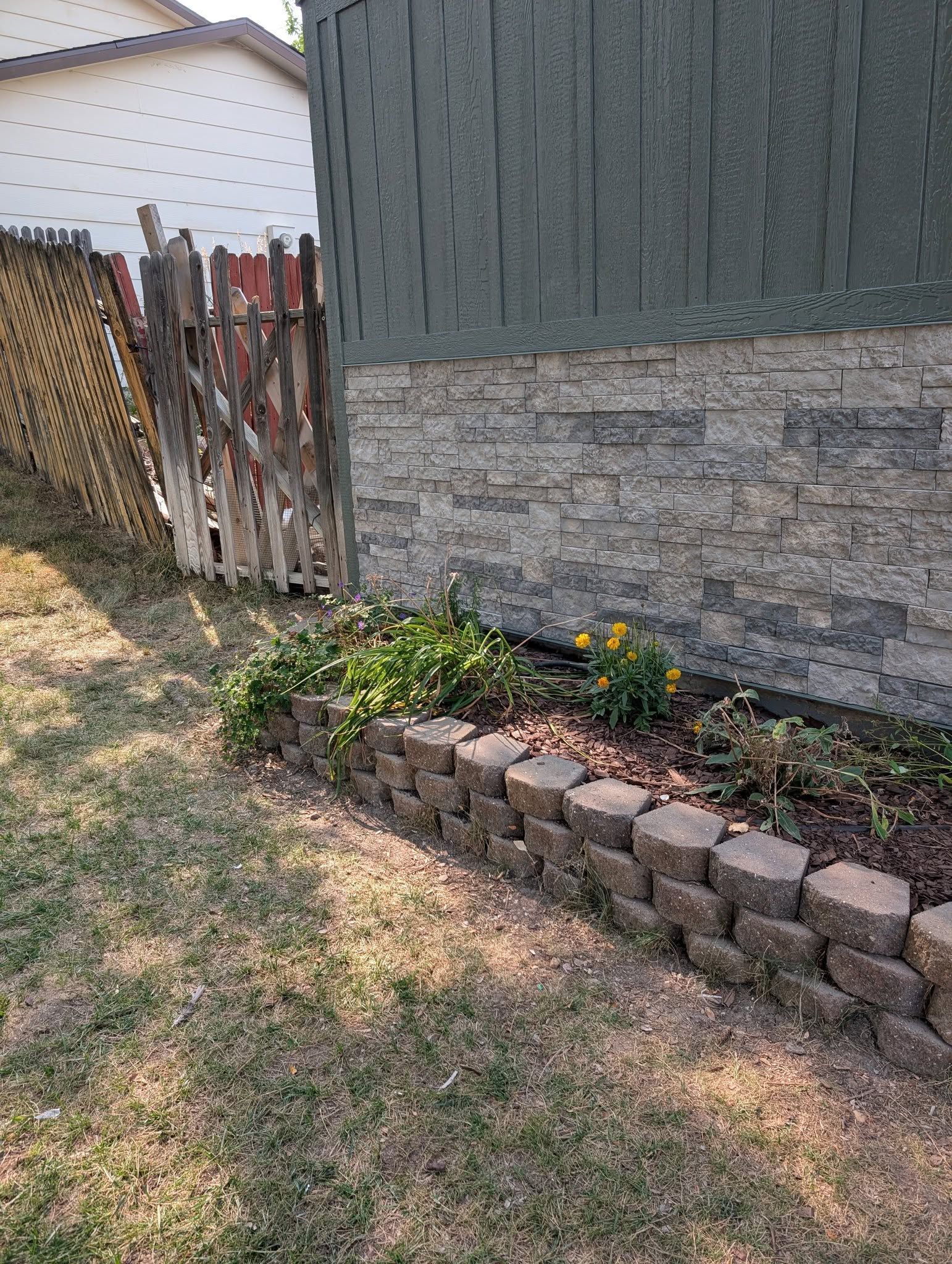 Backyard corner with a stone house wall, flower bed, and weathered wooden fence gate