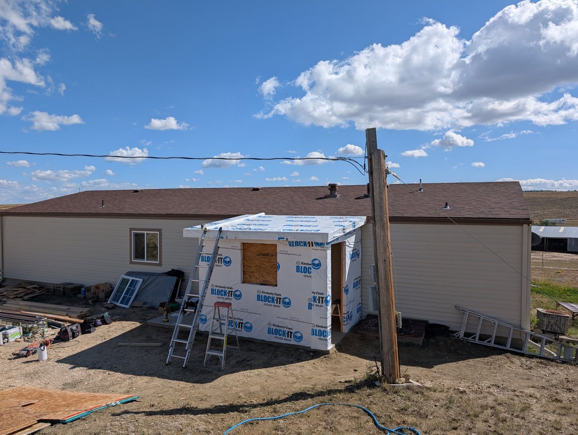 Partially built house with wrapped porch, ladder, and solar panels under a bright blue sky