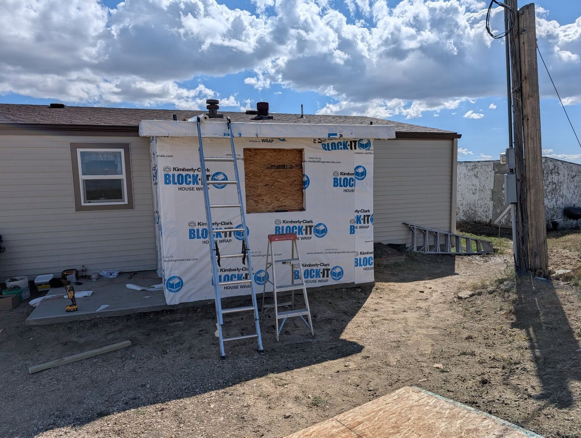 Damaged house exterior with boarded window, ladders, and protective wrap under a cloudy sky