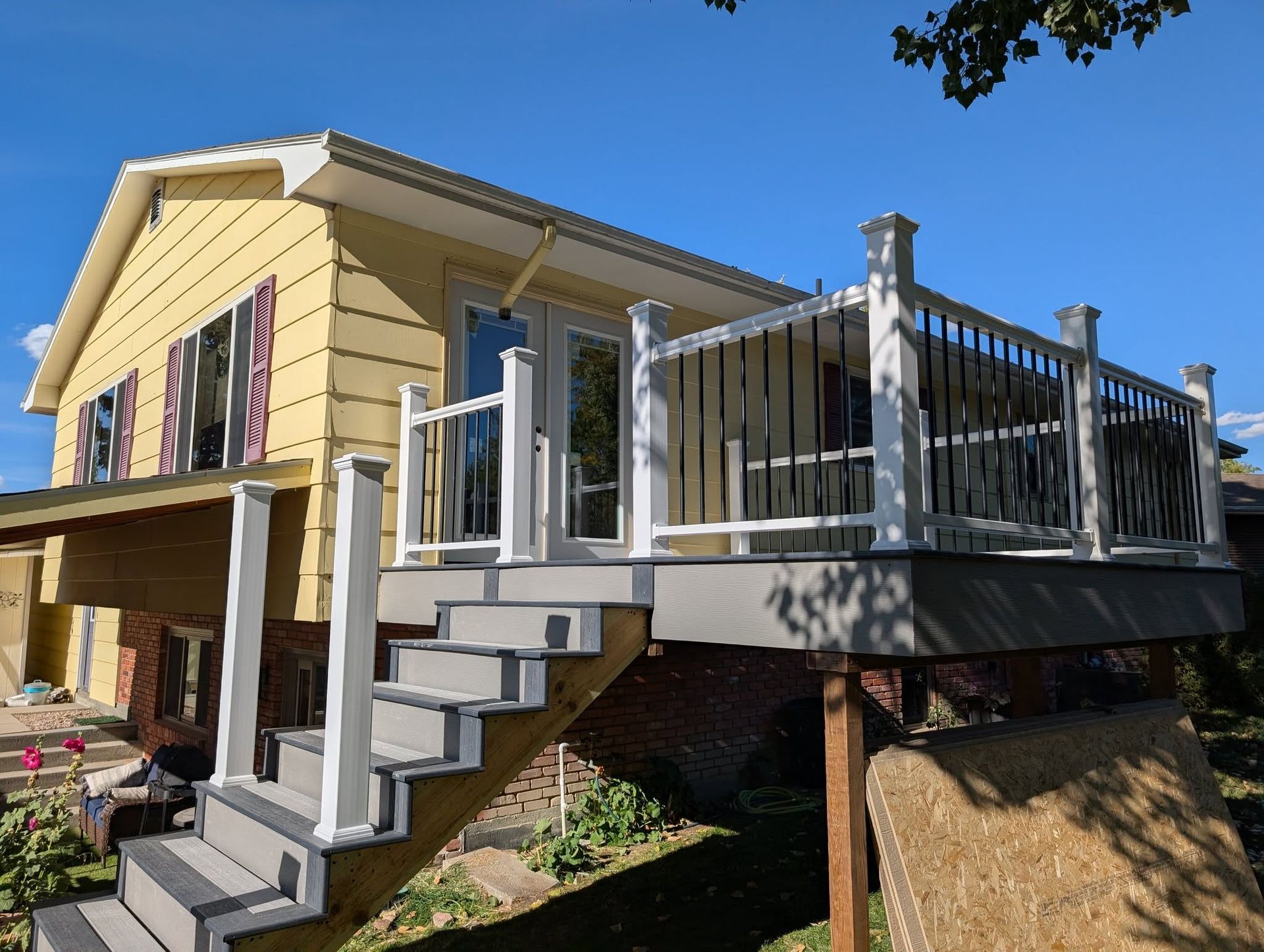 Yellow house with raised deck and stairs, white railing, and a brick foundation in a sunny yard
