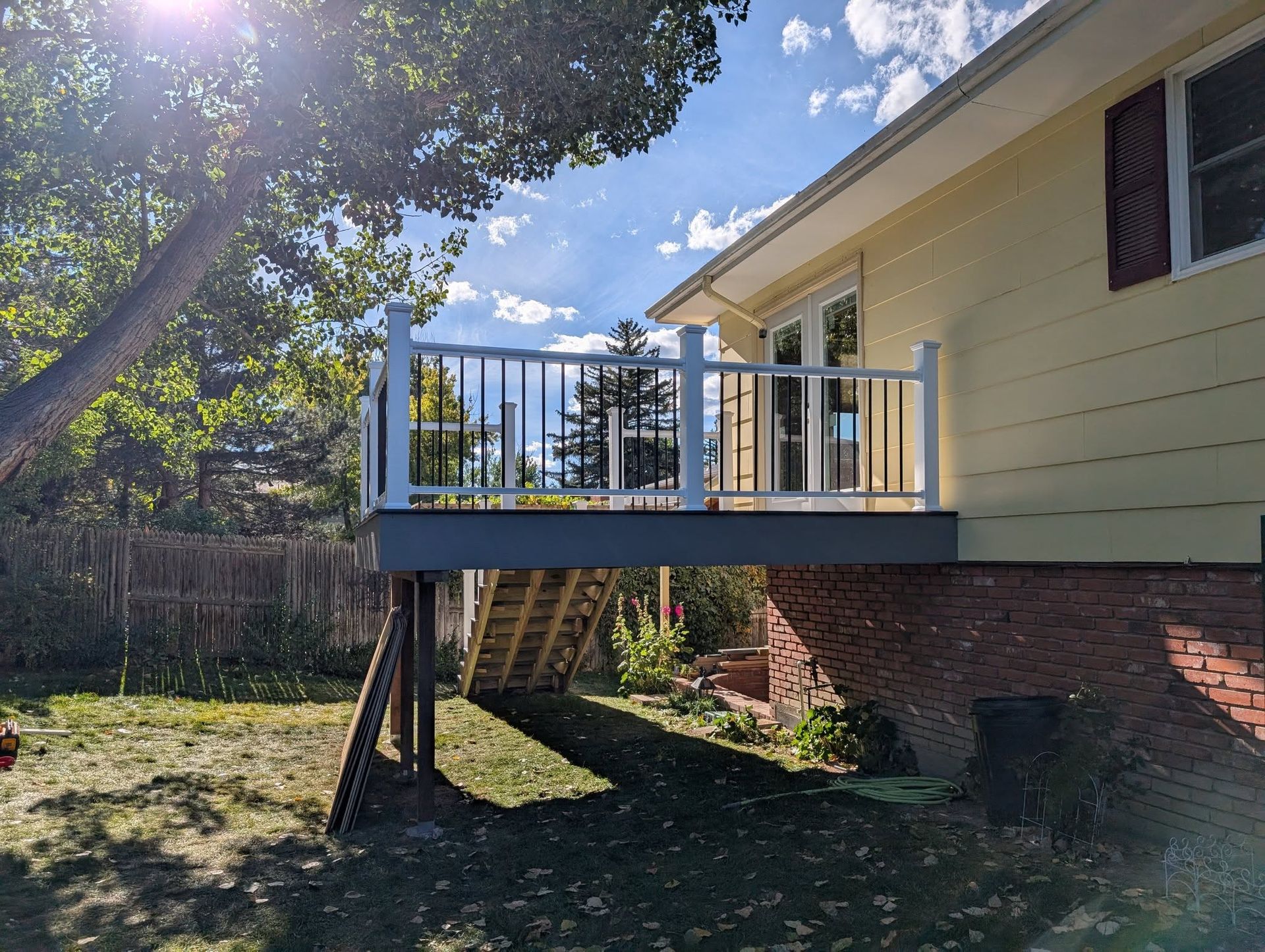 Raised wooden deck attached to a yellow house, with stairs down to a grassy yard and brick foundation.