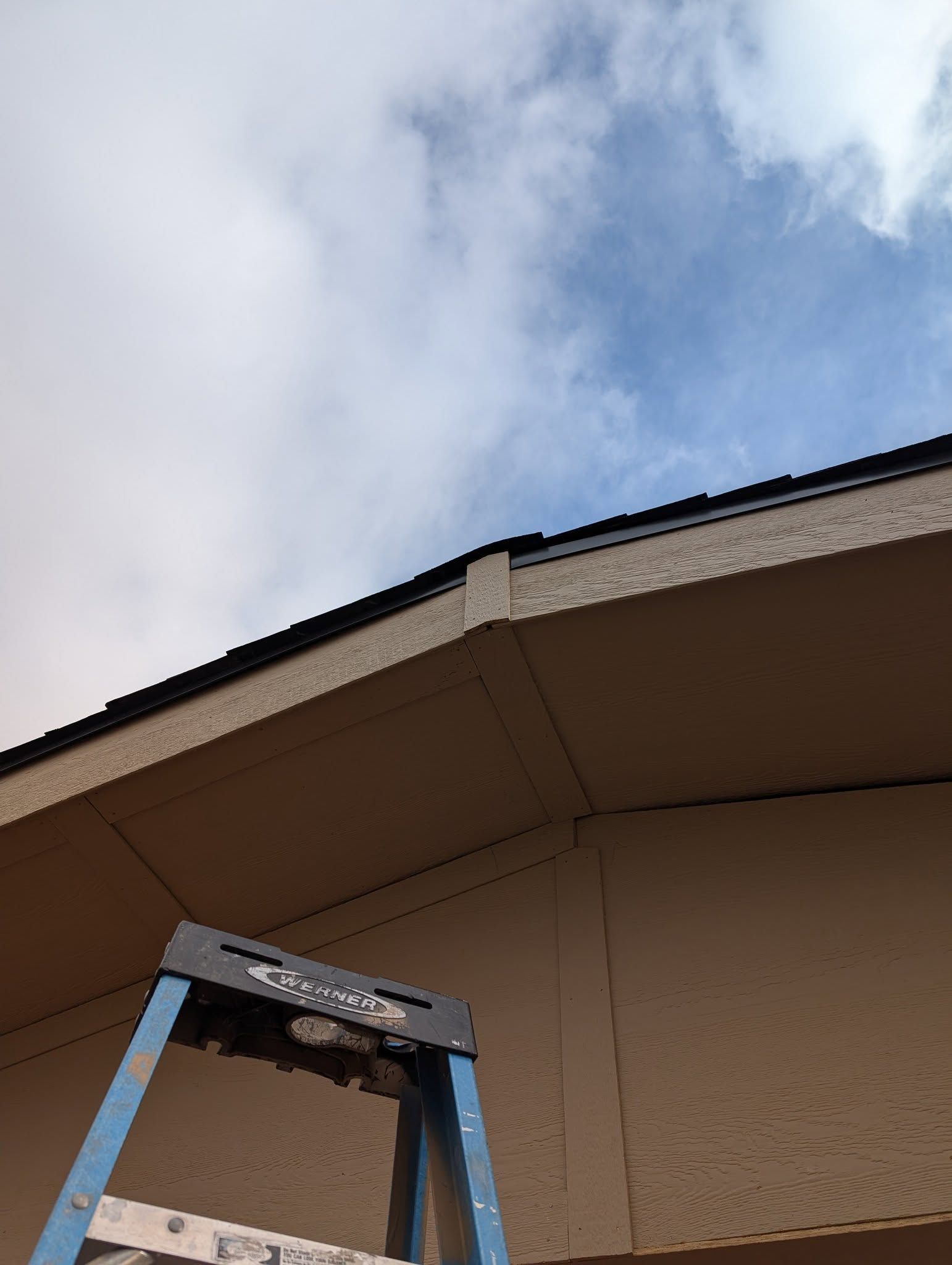 Blue ladder beneath a tan house roof against a cloudy sky
