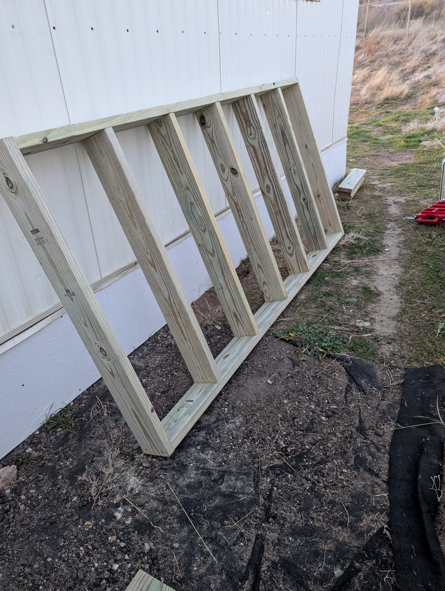 Wooden frame leaning against a white wall beside a gravel path and dry grass.